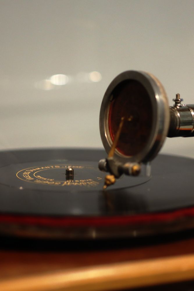 A close-up photograph of a record disc on a pathéphone (a French version of the phonograph/gramophone) from the 1910s. The photo shows the black disc lying on a red cloth turntable, with a thick metal arm ending in something resembling a large metal wheel holding a small needle down against the record. The record has some golden-yellow text in French at the centre, largely illegible due to the angle and the narrow depth of field. Part of it could be ‘Valse des Diamants’ – The Diamond Waltz.