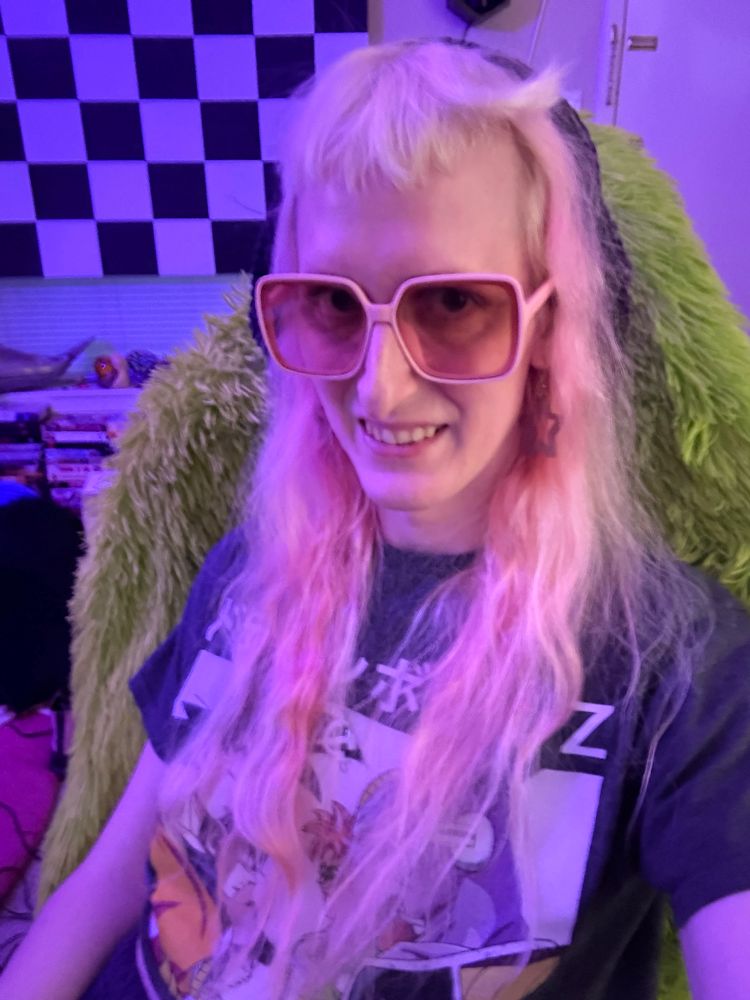Gwynn sitting in her computer chair smiling wearing a black beret pink rose tinted glasses, a hammer and sickle necklace a dragon, Ball, Z crop top and star earrings