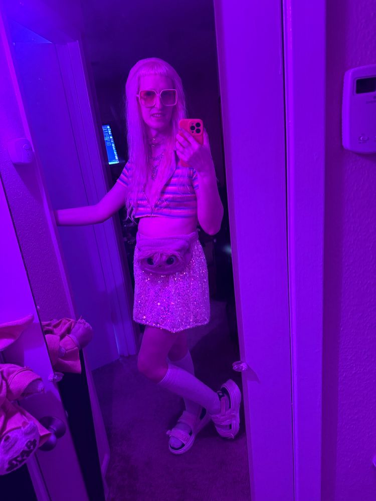 Gwynn full length mirror pic she is wearing a pink beret, pink rose tinted glasses, a pink collar, various necklaces, a stripped pastel crop top unzipped to the bottom but still covering boobs, a white tine stone skirt, white knee socks, and white and pink converse sandals