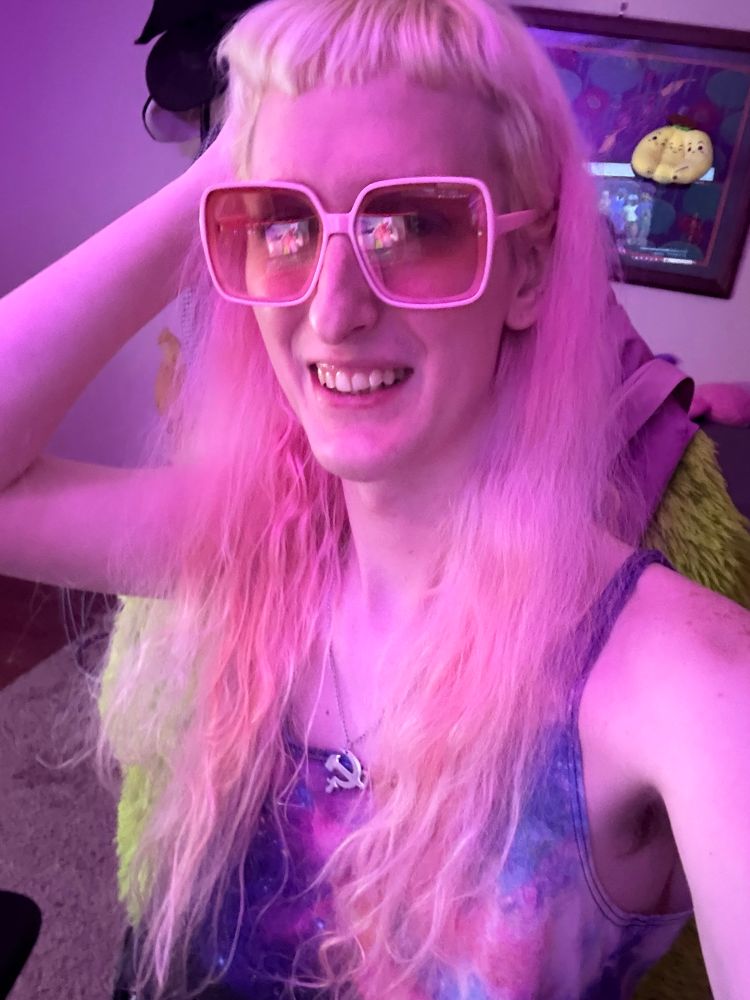 Gwynn sitting in her computer chair smiling, she is wearing pink rose tinted glasses. A hammer and sickle necklace, and a galaxy crop top