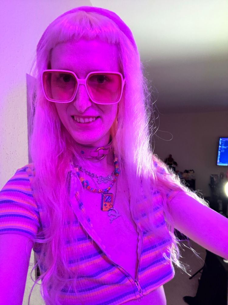 Gwynn selfie she is wearing a pink beret, pink rose tinted glasses, a pink collar, various necklaces, a stripped pastel crop top unzipped to the bottom but still covering boobs