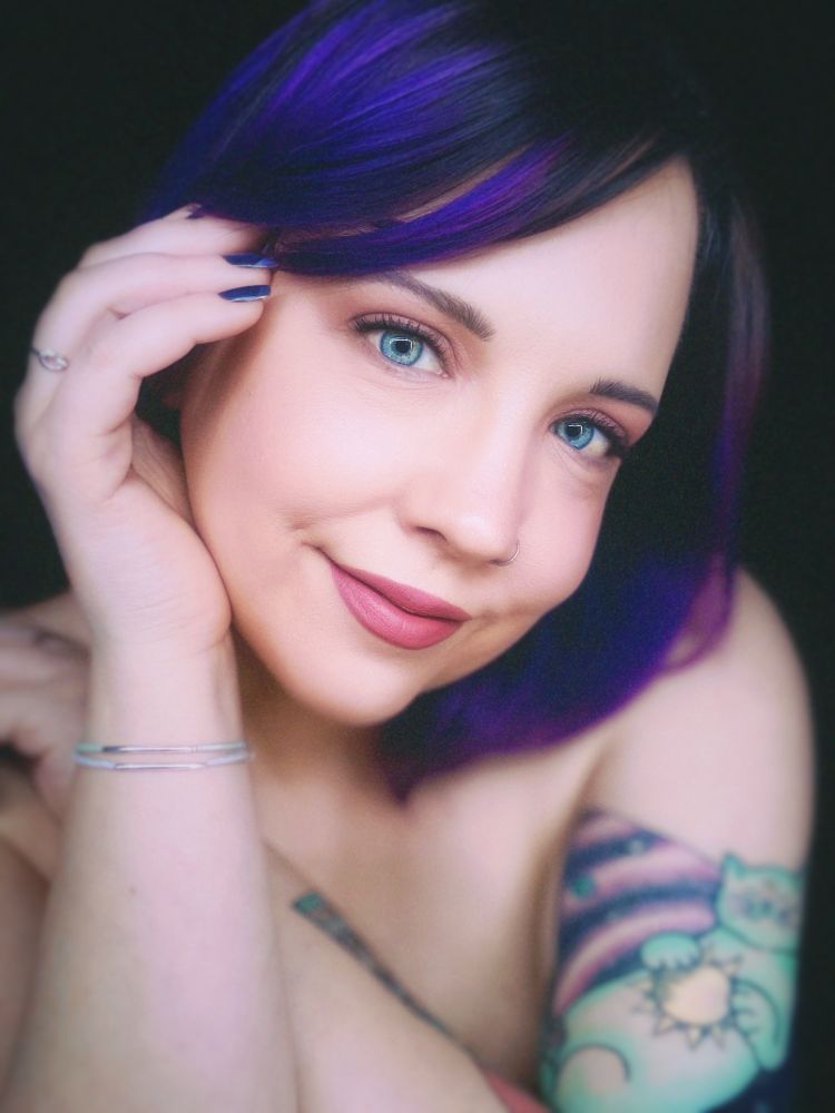 A woman with indigo hair is smiling at the camera with her hand softly pushing her hair back. She is topless and has a tattoo of a cat in a galaxy on her upper arm. 