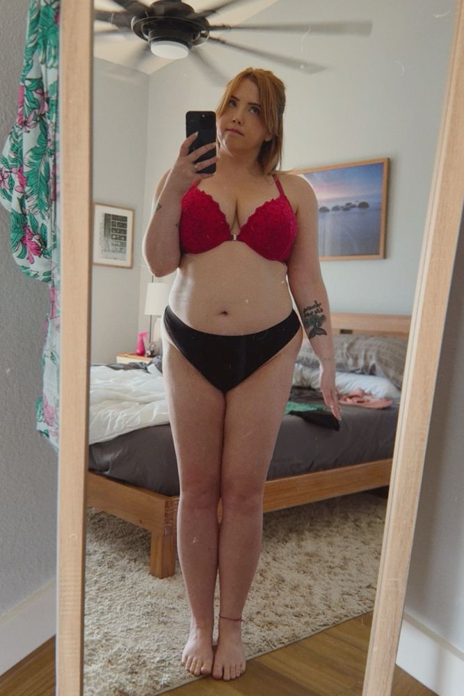 Cat is standing in front of a full length mirror in a pink push up bra and black thong. She is standing stiff with her arms by her side and making a quizzical face. 