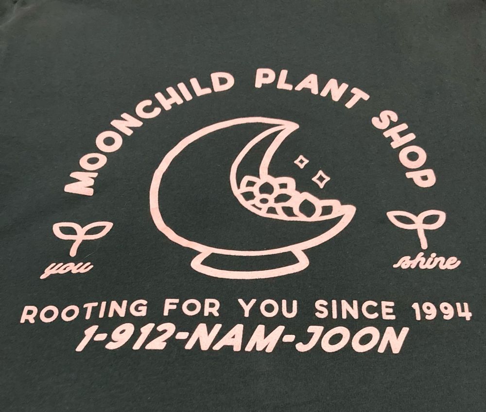 Graphic t-shirt with an open-moon-shaped planter and plant in the center. Arced above the words say “Moonchild Plant Shop.”  Below it says “Rooting for you since 1994.”  Below that it says “1-912-NAM-JOON.” On each side of the planter are two small saplings. In cursive script under the left sapling is the word “you” and under the right sapling is the word “shine.”