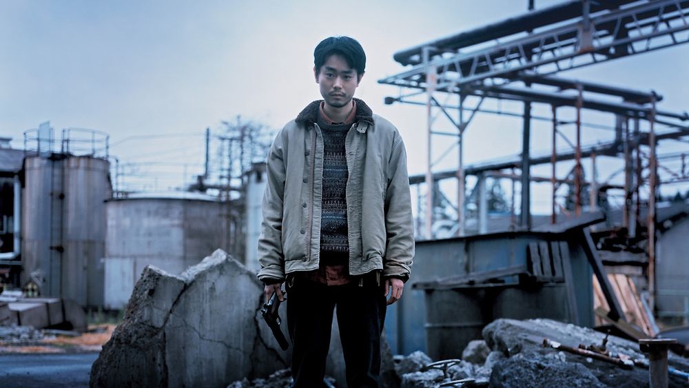Lead Masaki Suda looks directly at the camera, standing in an abandoned factory, a gun in his hand like an afterthought.
