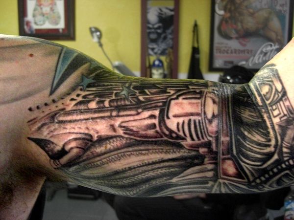A cybernetic style tattoo covering my left shoulder, bicep, and inner elbow, with the interior yellow wall of a tattoo parlor behind me