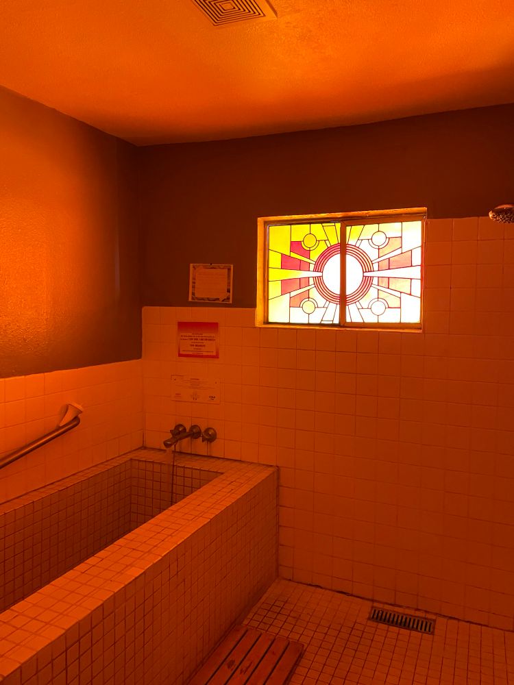 A tiled soaking tub in a small spa room illuminated by a warm orange glow. The walls and floor are covered in white square tiles, and a stained glass window above the tub features a geometric sunburst design in red, orange, and yellow tones. A wooden bench sits on the tiled floor next to a wall-mounted grab bar and faucet fixtures.