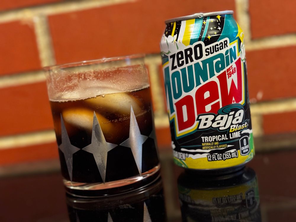 A high all glass with ice cubes and a brown liquid, next to a can of zero sugar Baja blast, in front of a red brick wall. 