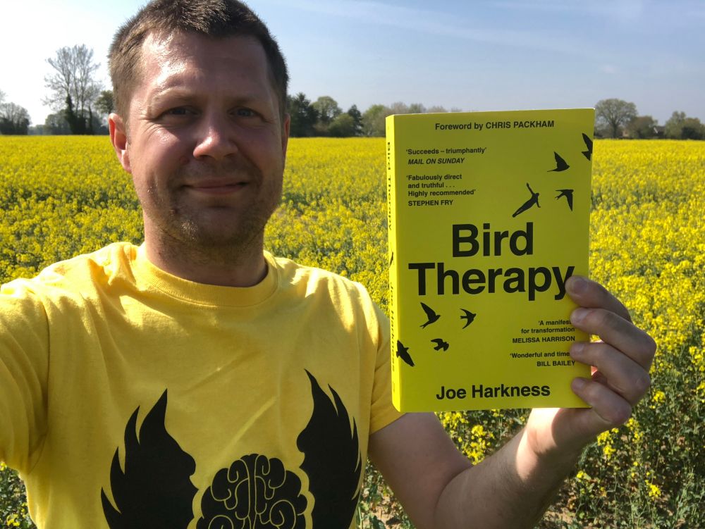 The author, Joe Harkness, stands to the left holding his book, Bird Therapy. He doesn’t look it, but he’s struggling with his depression on the day this is take . The book is bright yellow, he’s wearing a bright yellow y shirt, and is standing in front of a bright yellow field of oilseed. 