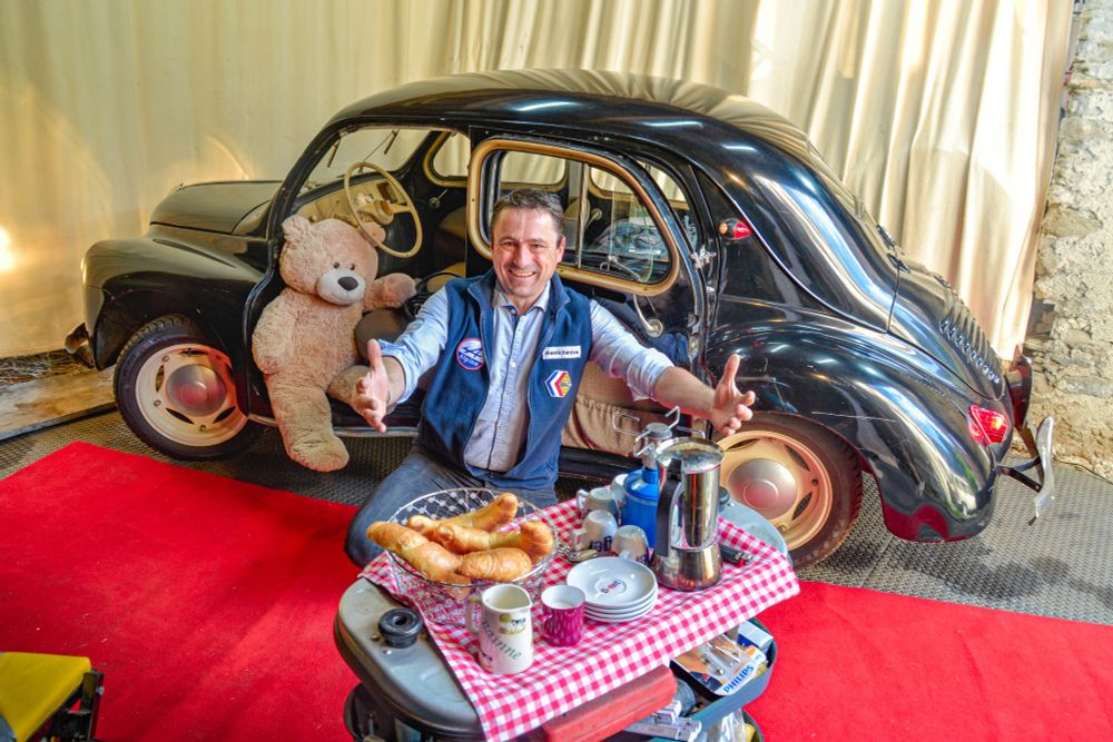 Remi zeigt auf Croissants und Kaffee auf einem Werkzeugwagen. Im Hintergrund ein schwarzer Oldtimer und ein Teddybär.