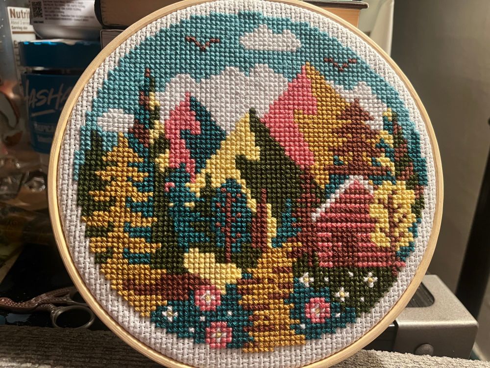 A completed cross stitch project showing a cloudy sky above a stylized mountain scene with multicolored trees, a walking path and a cabin. Pattern by Caterpillar Cross Stitch.