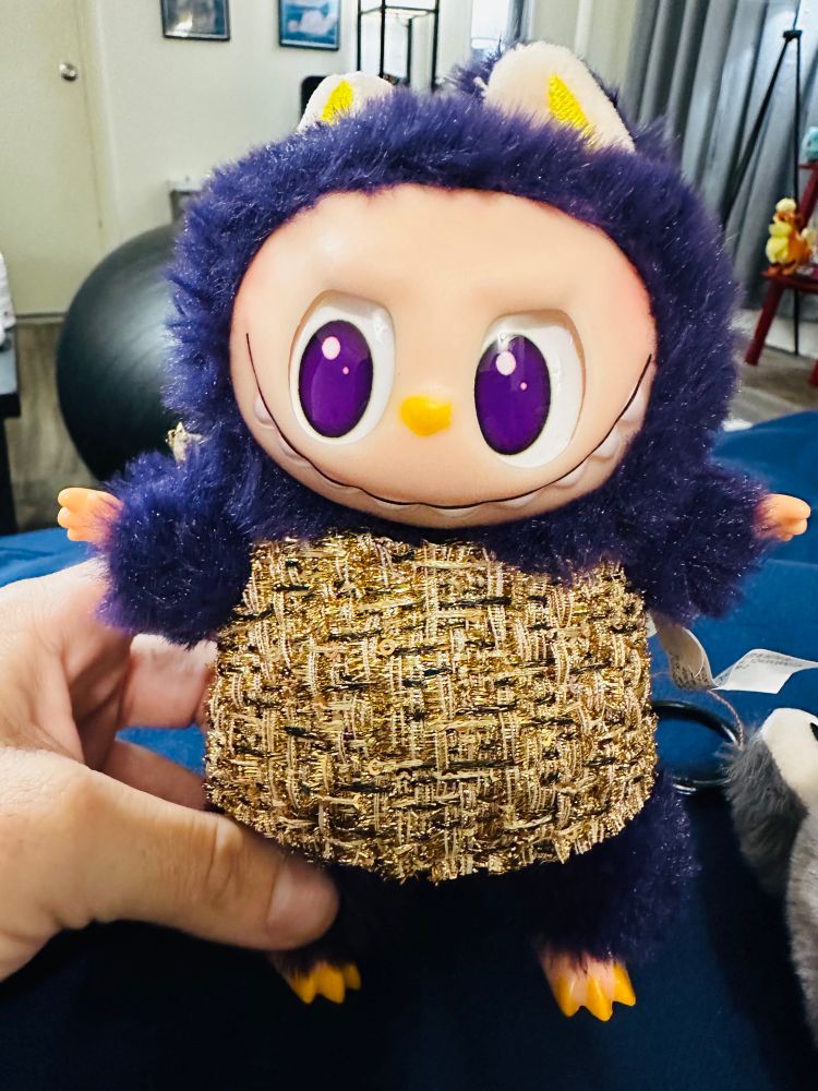 a hand holds a dark purple Labubu in a gold dress 