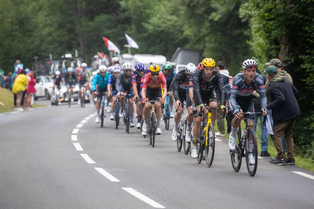 Eine Gruppe Rennradler bei der Tour den France fahren eine Straße entlang. Am Rand stehen einige ZuschauerInnen. Im Hintergrund ist Wald zu sehen.