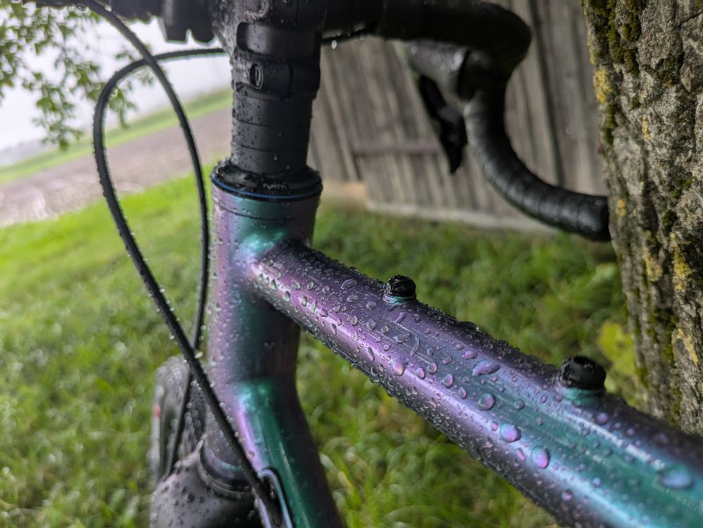 Ein Fahrrad Oberrohr von hinten fotografiert. Unten ist Rasen. Rechts ein Baum und eine Scheune. Auf dem Oberrohr sind Regentropfen. Der Lack ist Grün/Lila.