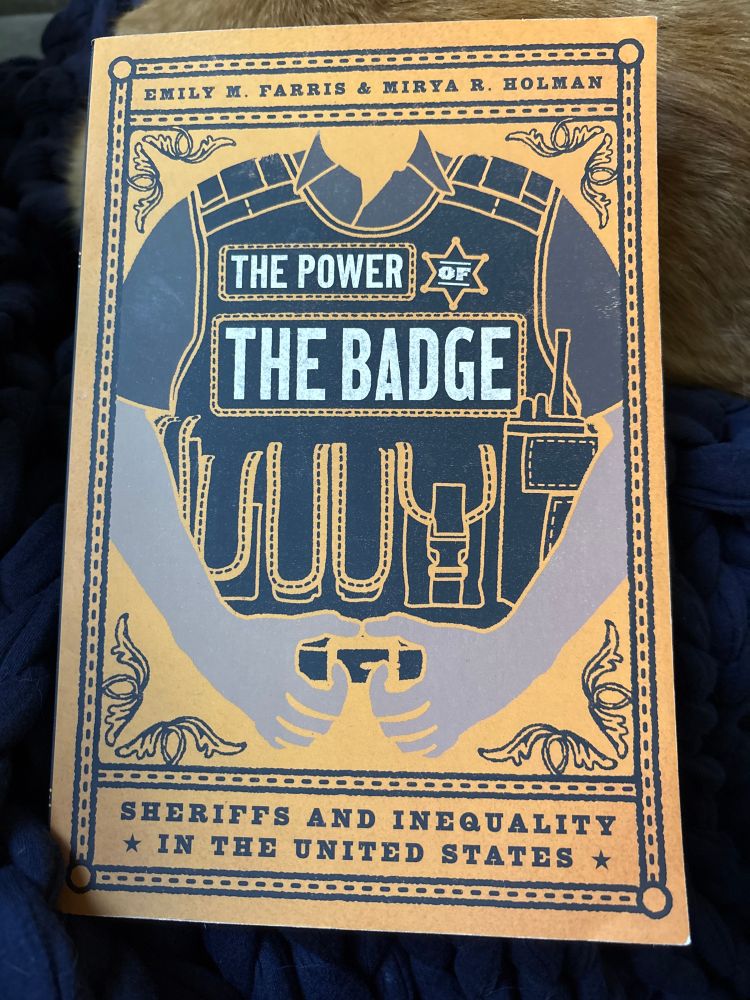 A picture of the book “The Power of the Badge” by Emily Farris and Mirya Holman. Seen in the background: Tonks, an oversized ginger house cat, allowing herself to be used as something to prop the book against.  