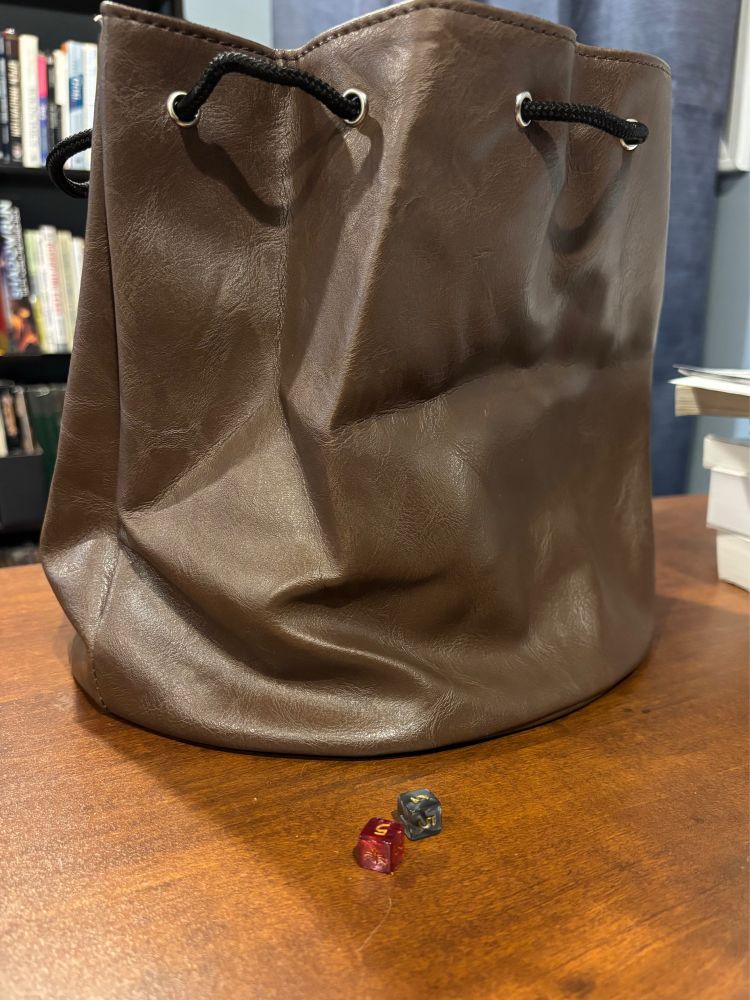 Brown leather dice bag with laced pull string. It has seven separate pockets inside (Unseen). In front, for scale, are 2d6, one reddish and one blueish. They’re mini dice from “Elderwood Academy” (a brand), so they make the already large bag look absolutely massive. Could probably stack 25 (or the mini dice) one atop the other and still not go from the wide bottom to the top of the bag. 