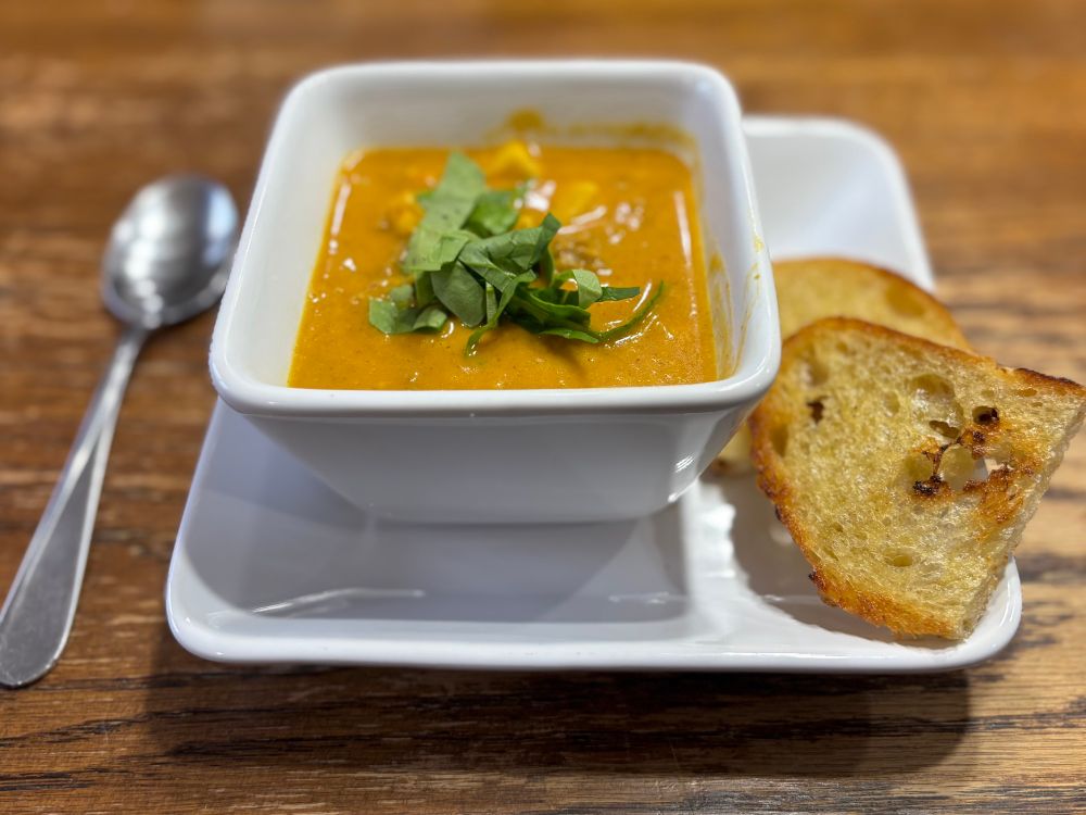 Both pictures of the same thing, from two different angles. A square bowl of Kathryn’s pumpkin curry soup on a square white plate. Topped with shredded spinach for garnish and paired with a pan-fried sourdough slice. 
