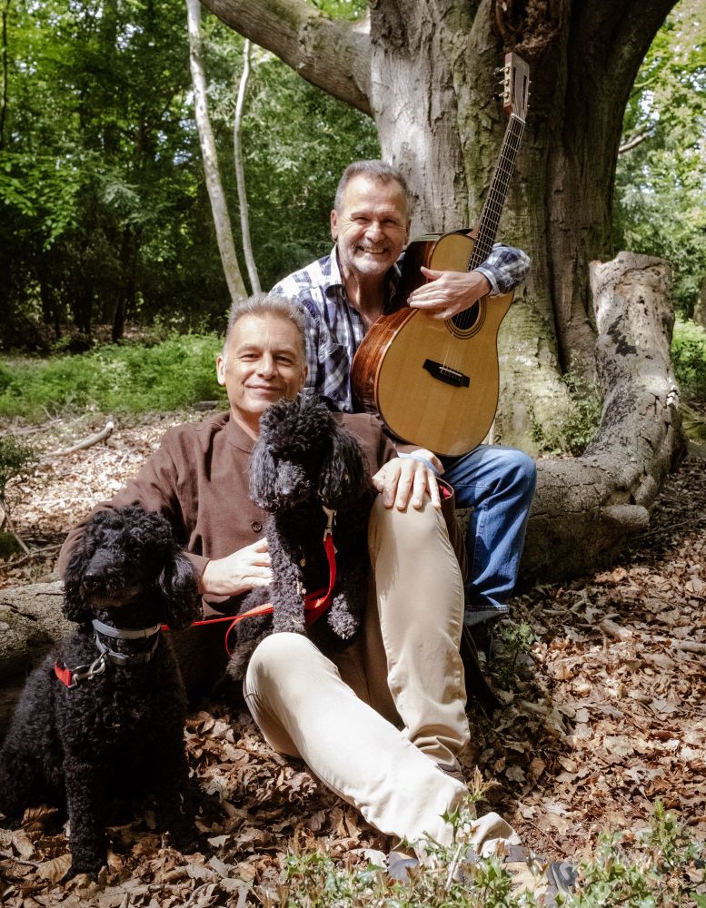 Chris Packham and Martin Simpson sat under a tree with Chris’s two lovely dogs, Sid and Nancy. Martin is holding his guitar and they both look relaxed and happy.