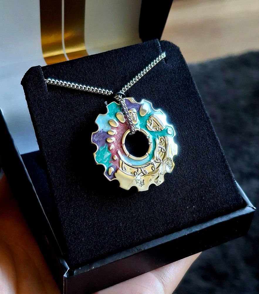 Image is a close up photo of the RockLove x Arcane collaboration Jayce and Viktor silver cog necklace with pearlescent inlay on the front facing side. It has swirling patterns of teal, purple, and rosy pink flooding the top left side of the cog, the unmarked parts shine in mirrored silver plate. End ID 