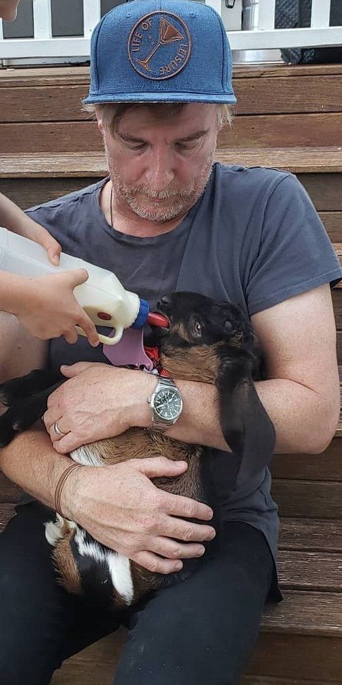 Rhys Darby holding a baby goat in his arms that's being bottle fed by someone off camera. 
