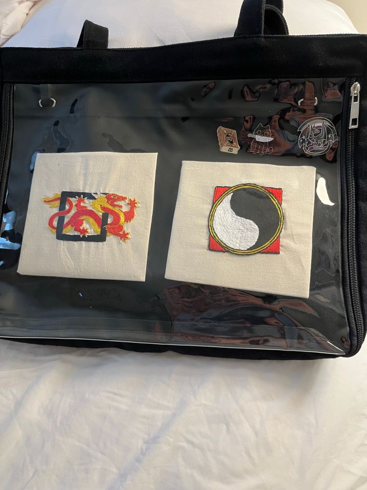 Black tote bag with clear vinyl front. Behind the vinyl are two squares of embroidery one the a red and gold dragon the other with a black and white circle. 