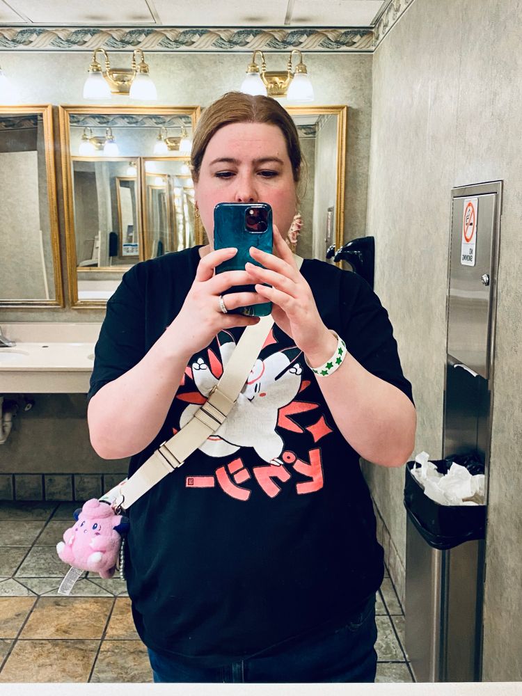 A mirror selfie of myself. I’m wearing a black t shirt with a clefable on it. A white purse strap goes me with a clefairy keychain on it. I’m hiding half my face behind my teal covered phone. Background is a bathroom.