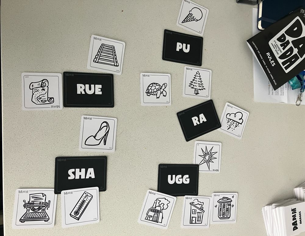 Black and white cards on a table. Black cards have white text with phonemes (PU RUE RA SHA UGG) and white cards have printed cartoon drawings of various items (e.g. a typewriter, trashcan, a turtle). The picture cards are grouped around the phonemes in a categorical way. The game rulebook is in the top right and says DADADA on the cover.