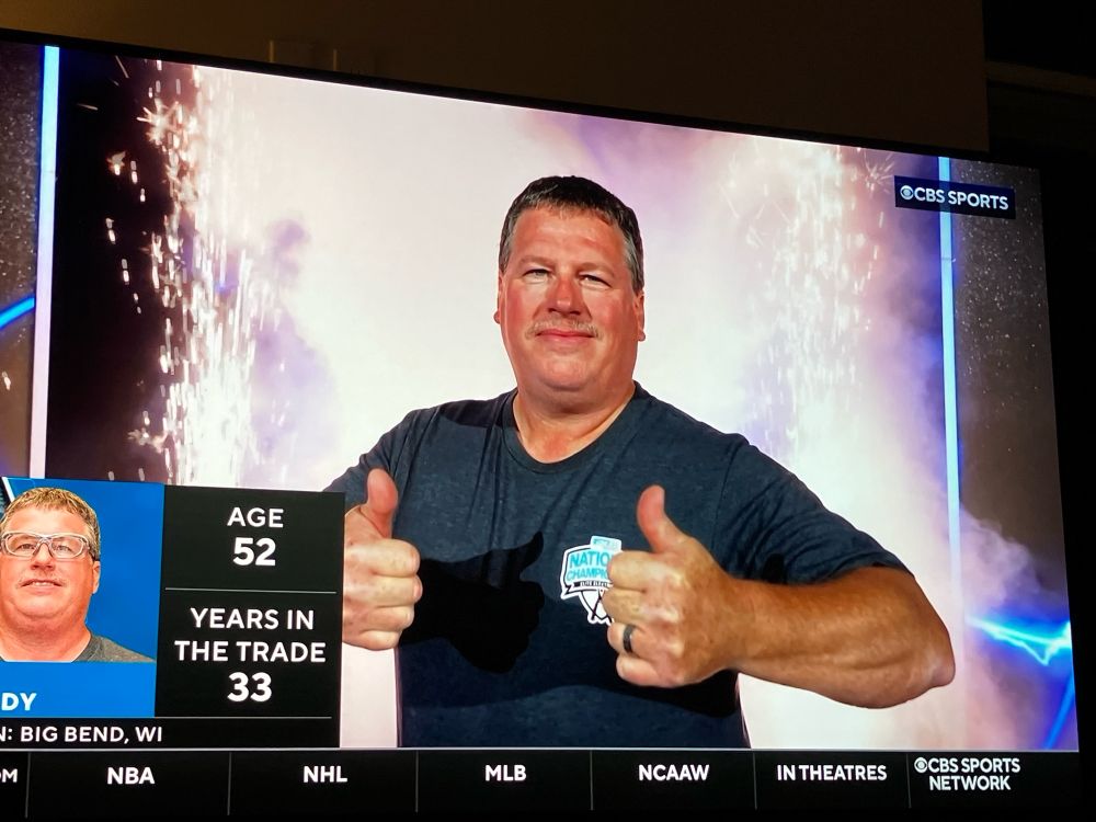 A contestant being introduced. A bug on the side says he’s 52 with 33 years in the trade.  He’s giving two thumbs up and there are fireworks going off behind him. 