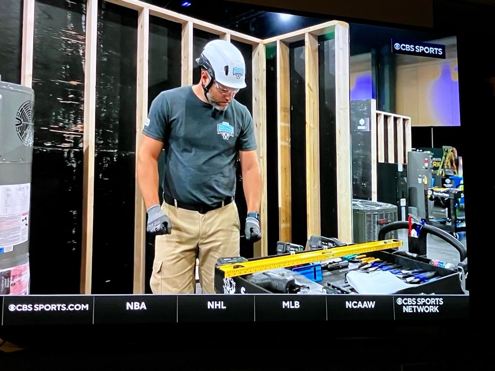 An electrician in a safety helmet and work gloves looking down at a tool shelf with a stripped wall behind him