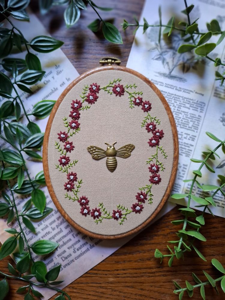 Broderie d'une couronne de fleurs rouges avec une breloque d'abeille attachée au centre. Tout cela sur une table en bois foncé avec des plantes et page de vieux livre en décoration 