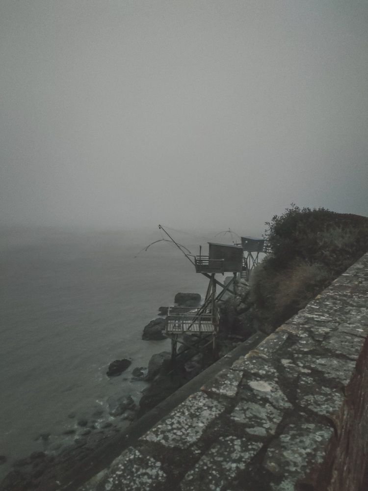 Photo de la mer avec du brouillard. En 1er plan on a un petit muret, ensuite 2 petites cabanes de pêcheurs. Et en fond la mer brumeuse.