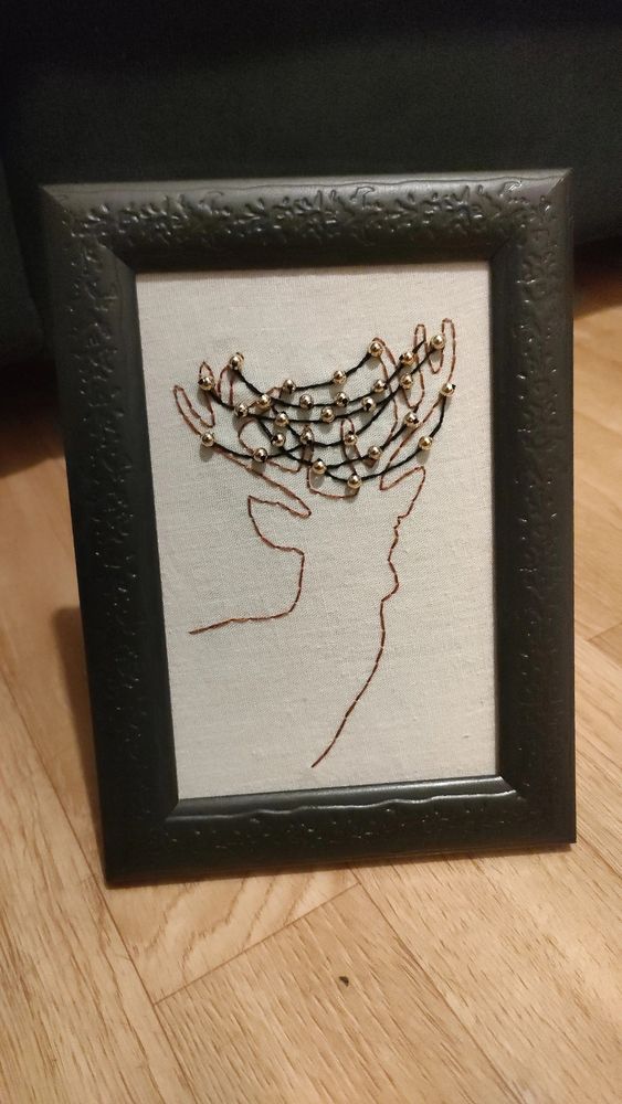 Broderie d'un renne de Noël qui a ses bois pris dans une guirlande de Noël avec des perles. Cette broderie est dans un cadre noir avec des petits détails style feuille