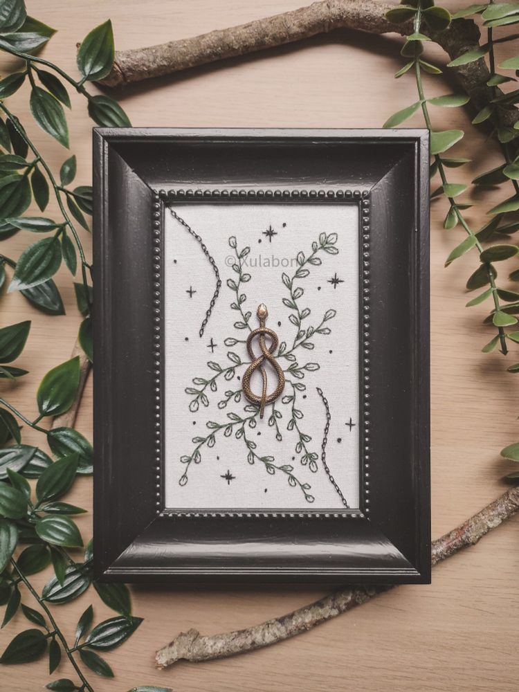Broderie d'un serpent attaché au centre avec du feuillage partant de celui-ci brodé ainsi que de petites étoiles. Des chaînes en haut a droite et bas gauche sortent du cadre.