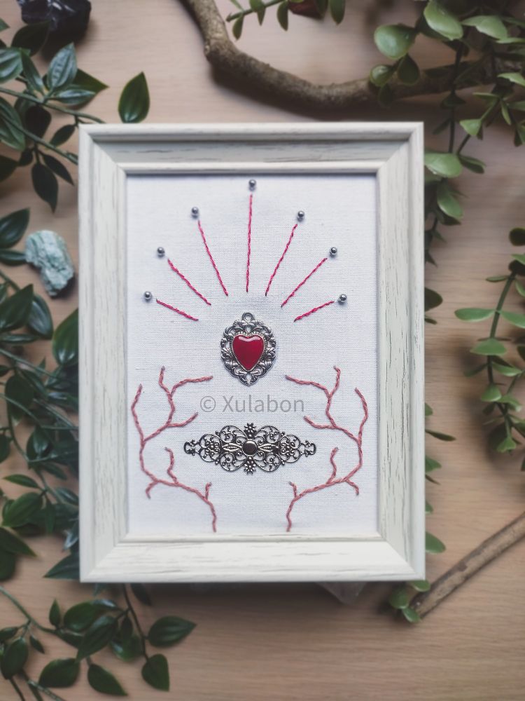 Cadre blanc avec un cœur en métal avec le centre rouge attaché au centre. Autour il y a des ornements de brodé et une piece ornementale attaché en dessous du cœur
