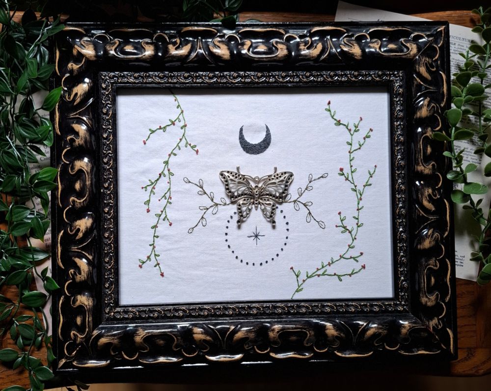 Broderie en cadre baroque avec un papillon en argent attaché au centre. Une lune brodée au-dessus, du feuillage autour et des ornements en dessous.