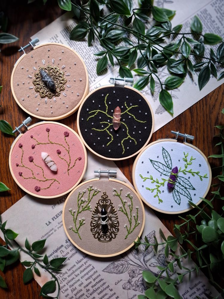 Photo de groupe avec les 5 broderies sur une table en bois foncé avec des pages de vieux livre en fond et des plantes vertes.