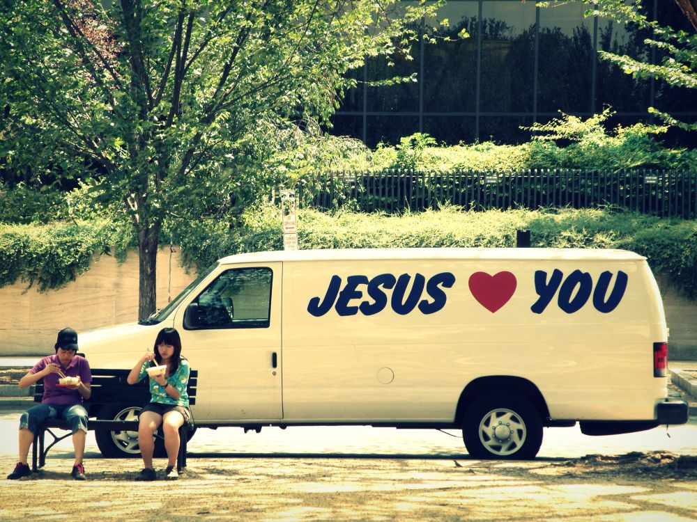 on the national mall: a couple sitting on a bench eating in front of a white van that says JESUS ❤️ YOU on the side
