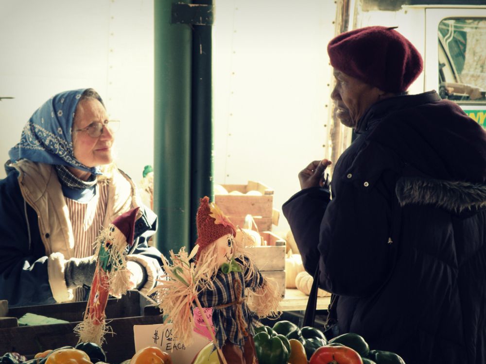 older lady in headscarf at eastern market at her booth talking with an older lady customer  in a great beret over a stand of vegetables