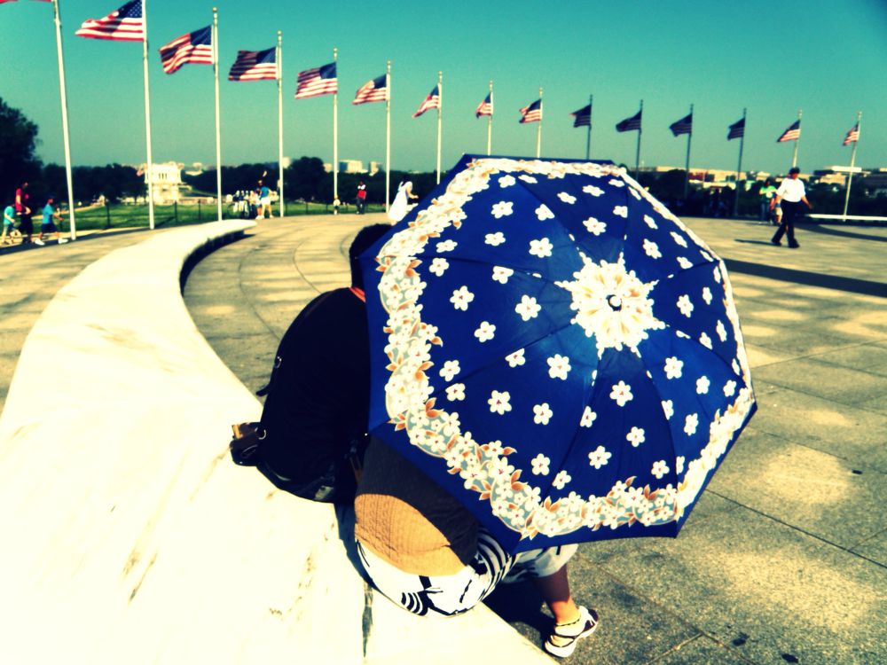 looking down on of the benches at the base of the washinton monument: background is sky and part of the ring of flagpoles; foreground is the top of an umbrella shielding a couple from the heat