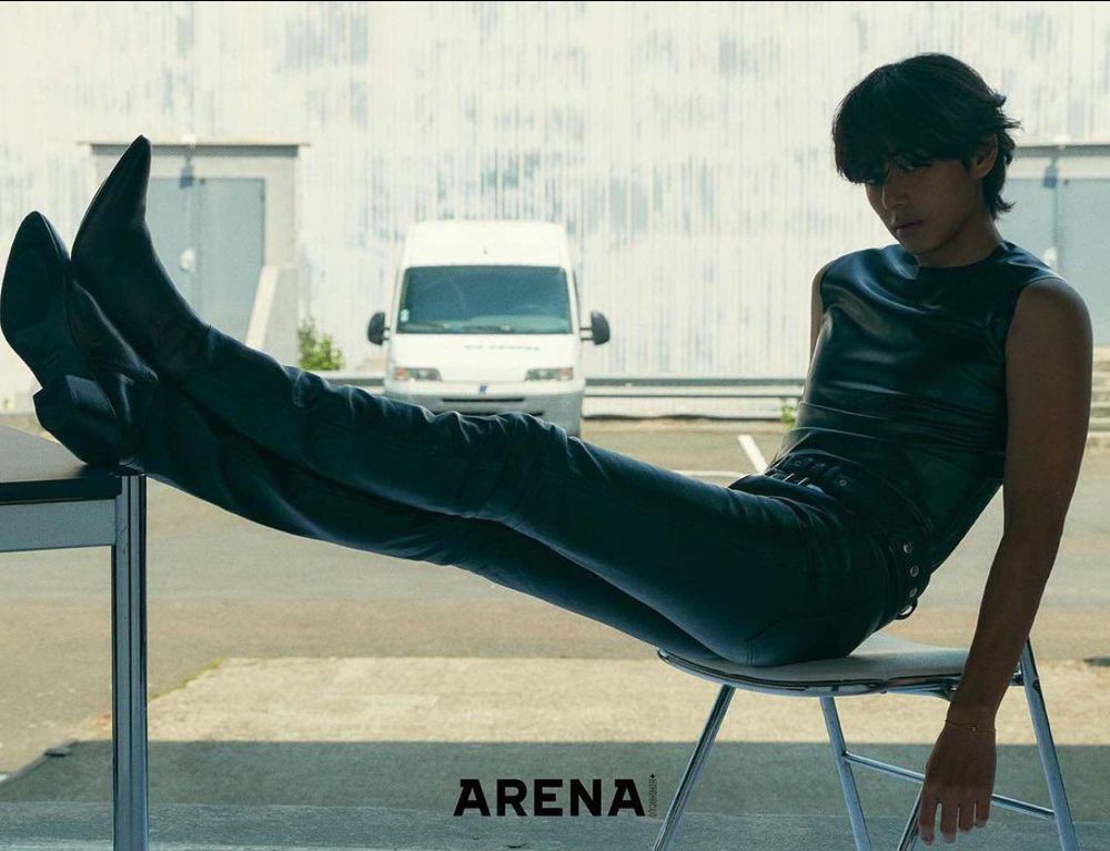 Taehyung of bts. He is sitting on a chair with his legs stretched out resting on a table edge. He has on a sleeveless shirt tight leather pants and heeled boots. He looks 90 percent legs in this picture.  