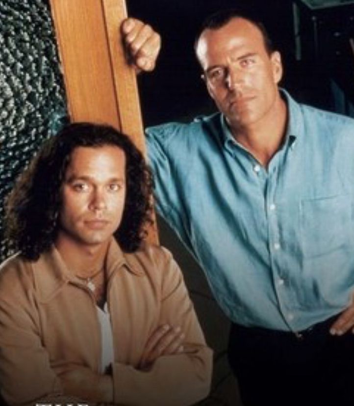 A pic of Blair Sandburg (played by garret maggart) and Jim Ellison (Richard burgi) from the 1996 show “the sentinel “ Blair has should length brown curly hair, a tan jacket and white shirt and is looking  into the camera with his arms crossed. Jim has very short  brown hair and blue button down shirt. It’s all so mid ‘90s it hurts. Jim is looking quite a bit bigger than Blair in this shot which really helped set us up for years of size difference kink in fanfic. *Decades* even. 