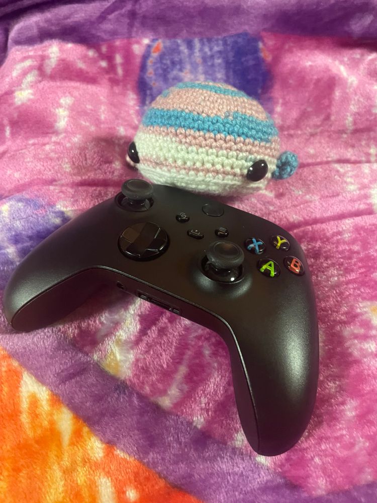 Close up of a hand crocheted whale in trans pride flag colors on top of an Xbox controller for scale against a very colorful throw blanket. 