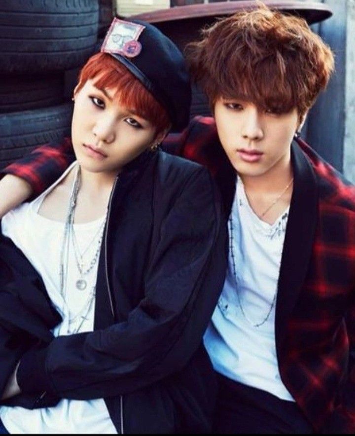 Min yoongi of BTS sitting on Kim seokjin of bts. Yoongi is wearing a black beret, white shirt and black jacket and several silver chain necklaces. With red hair and heavy eyeliner he is looking right into the camera with a “you want me but he has me.” Thing going on.  Jin has chestnut hair and is wearing a white shirt under a red and black plaid jacket and few silver necklaces. His arm is around yoongi’s shoulders and his eyeliner is also saying a lot. 