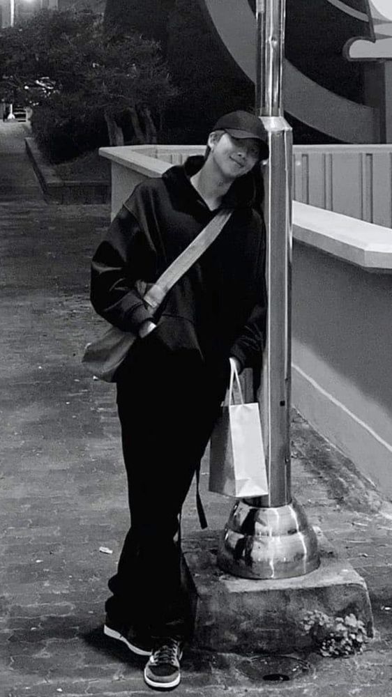 Namjoon of bts is leaning against a lamppost looking into the camera. It’s a black and white pic, he is wearing long pants, a hoodie, a baseball cap and a cross body bag holding a shopping bag and smiling the most gentle smile. The most boyfriend pic ever. EVER! 