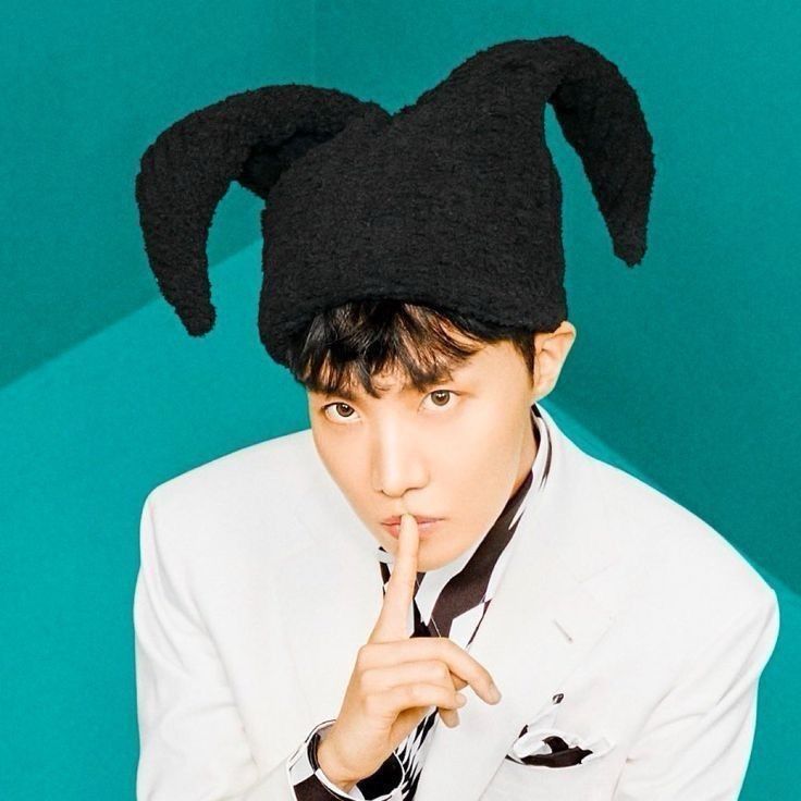 Jung Hoseok of bts in concept photos from his jack in the box album. He is in a white suit with a black and white shirt and a black jester hat. He is holding his index finger to his lips in a shush pose against a teal background.