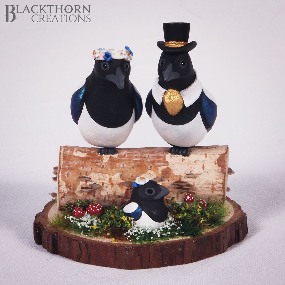 Two Magpies sit upon a log with fungi on it. One has a braided headband with blue and white flowers on it and the other has a black top hat with a golden band and a matching cravat. Sitting in the grass is a small Magpie chick with flowers on it's head.