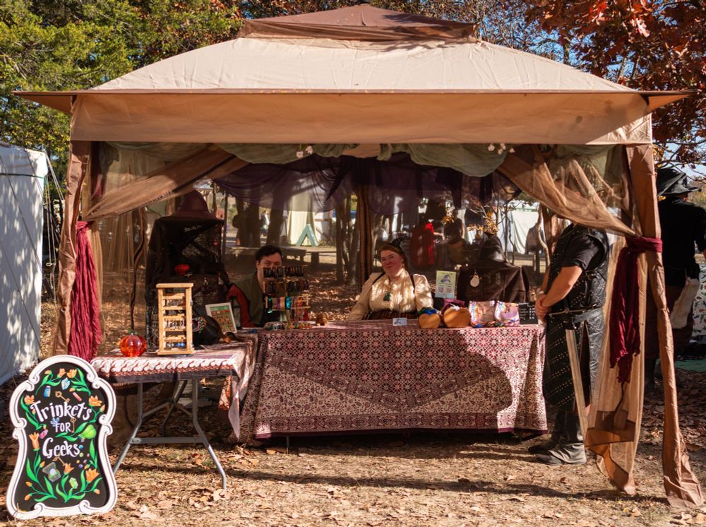 A tan and brown canopy shades tables covered in various wares with a sign that reads “Trinkets for Geeks” to the side. The vender and their assistant sit behind the tables in ren faire garb while a patron stands to the side.