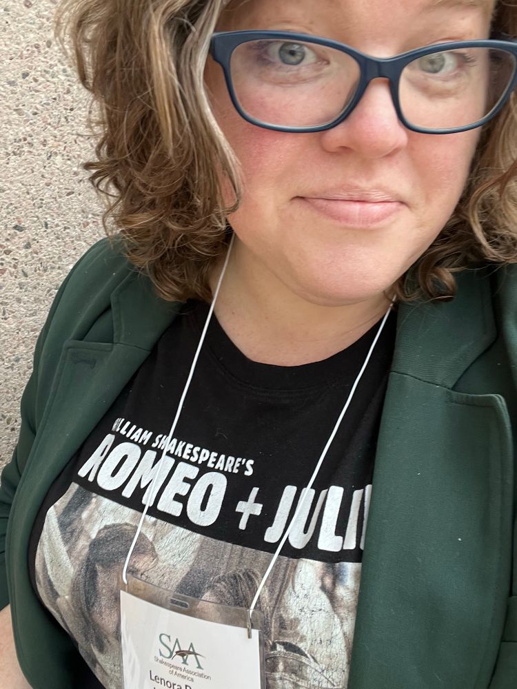 Selfie of a white woman with curly brown hair wearing blue glasses, a green blazer, and a black t-shirt with a graphic from Baz Luhrmann’s ROMEO + JULIET. SAA name tag partially obscures shirt. 