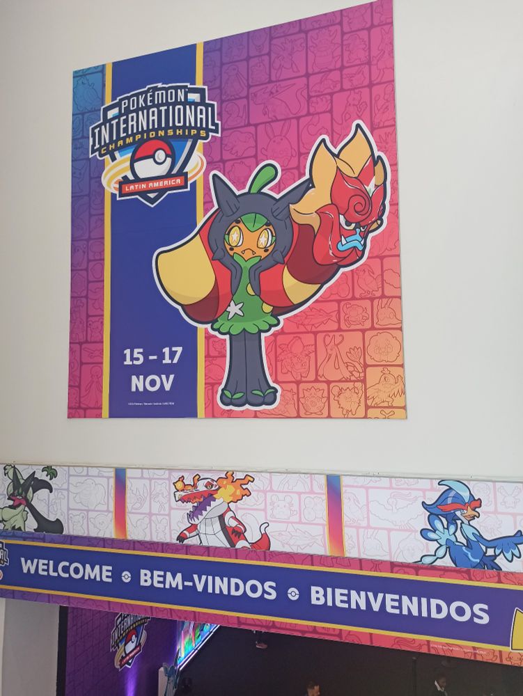 Decoração acima da entrada do evento, com os dizeres "bem vindos" em português, inglês e espanhol, adornado com os pokémon inicial de Paldea na última forma: Meowscarada, Skeledirge, e Quaquaval.
Acima ainda outro poster com a logo do evento, as datas (15 - 17 de novembro) e a Pokémon Ogerpom com sua máscara Hearthflame