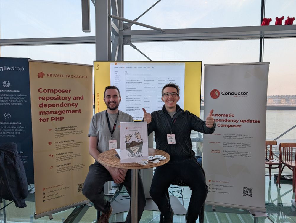 Two people on stools at a table in front of a Private Packagist and a Conductor banner as well as a big screen.
